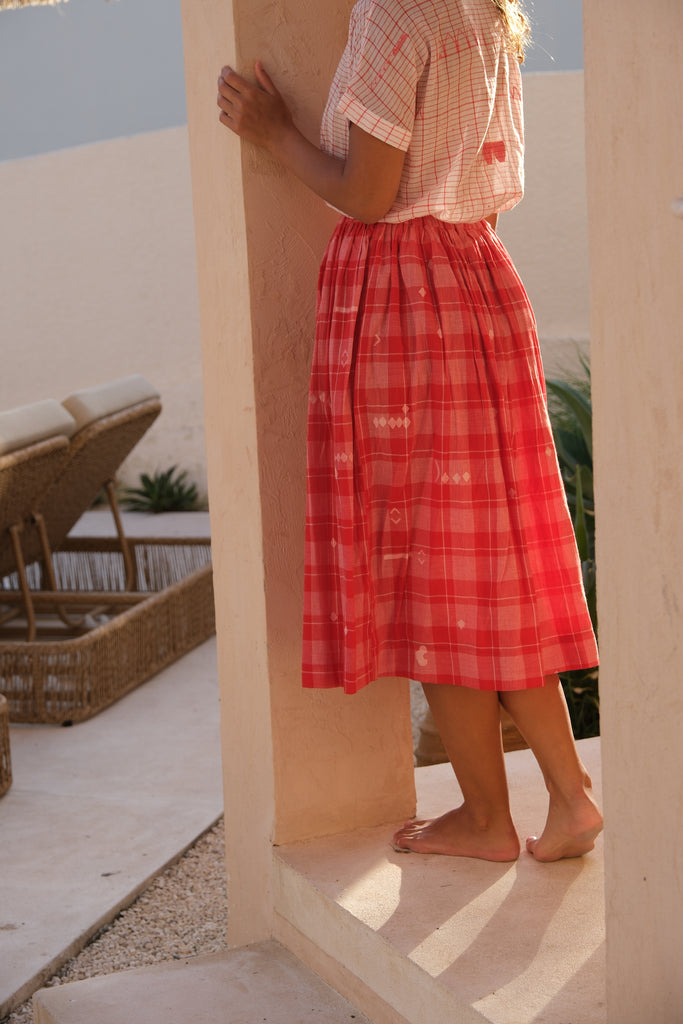 Introducing the Crimson Checkered Cuban Shirt, a collaboration between Audrey E Leary and World of Crow. This hand-woven jamdani fabric shirt is light pink with red motifs, short sleeves, and is super light and breathable, perfect for everyday wear. Sustainably made from soft cotton, paired with a red checkered skirt.