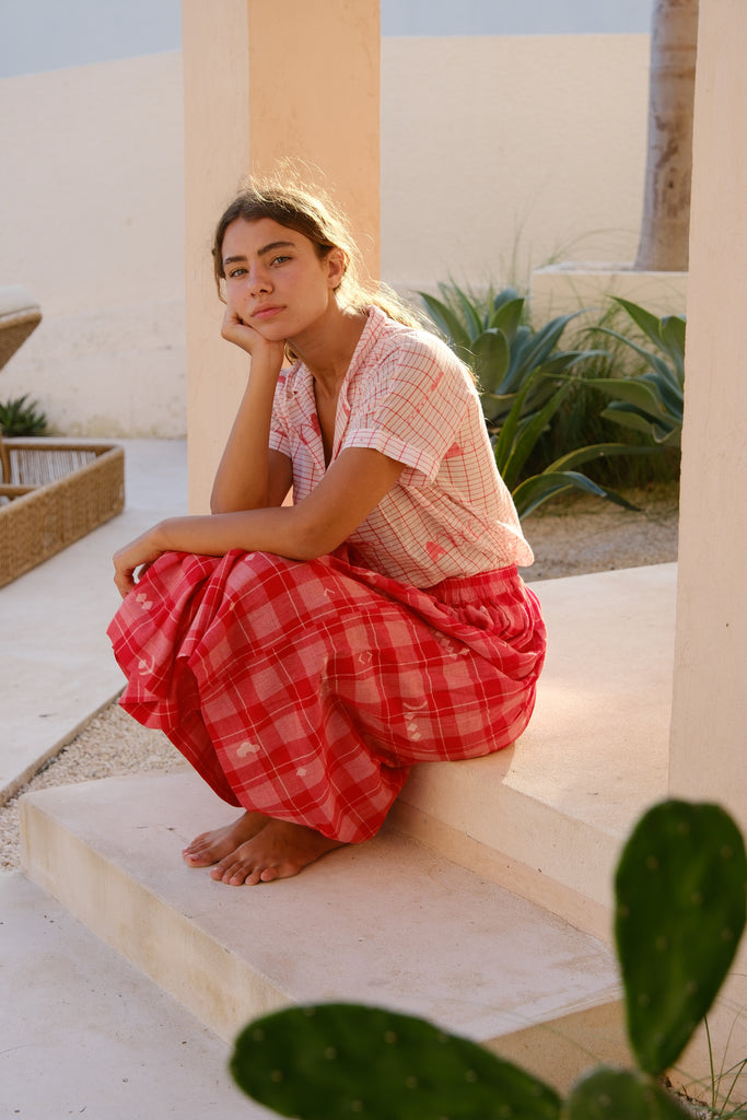 Introducing the Crimson Checkered Cuban Shirt, a collaboration between Audrey E Leary and World of Crow. This hand-woven jamdani fabric shirt is light pink with red motifs, short sleeves, and is super light and breathable, perfect for everyday wear. Sustainably made from soft cotton, paired with a red checkered skirt.