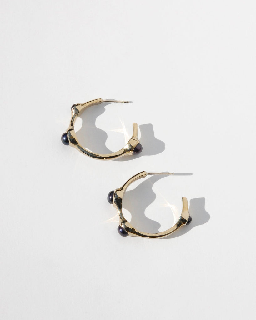 Introducing our Gold coloured Hoop Earrings adorned with three striking black pearls. Crafted in solid brass or sterling silver, these elegant earrings feature a slightly asymmetrical design, with a refined 1 1/4" diameter. Perfect for making a sophisticated statement, they blend timeless elegance with a touch of modern flair.