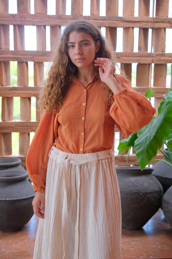 Introducing the Audrey X Crow Collection, featuring a woman in an orange Jane Austen Shirt and white skirt. This collection showcases hand-woven fabric with block-printed stripes, embodying elegance and artisanal craftsmanship. The vibrant orange and classic white combination adds a touch of sophistication to this beautifully crafted ensemble.