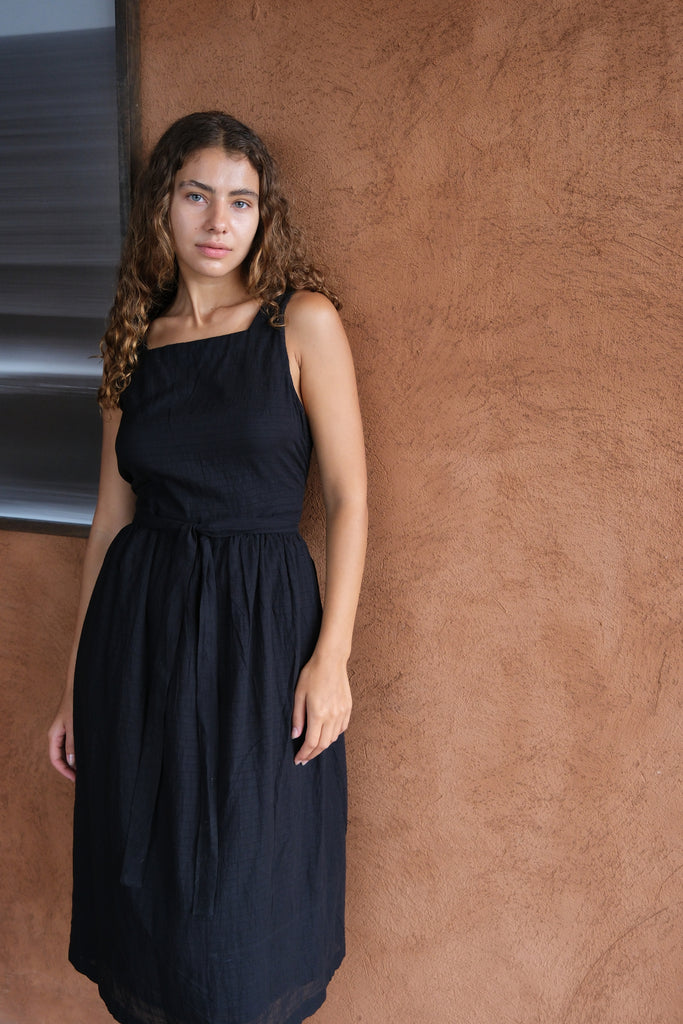 Introducing The Classic Hitch Dress, a collaboration between Audrey E Leary and World of Crow. This black midi-length dress features a square neck and a wrap-on design with a drawstring at the waist. Sleeveless with a straight hem, it is crafted from soft, lightweight fabric that offers a comfortable fit over the bust and a relaxed fit around the body. True to size, this dress combines elegance with ease for any occasion.