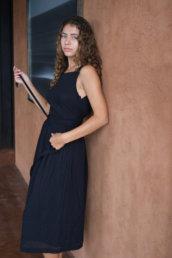 Introducing The Classic Hitch Dress, a collaboration between Audrey E Leary and World of Crow. This black midi-length dress features a square neck and a wrap-on design with a drawstring at the waist. Sleeveless with a straight hem, it is crafted from soft, lightweight fabric that offers a comfortable fit over the bust and a relaxed fit around the body. True to size, this dress combines elegance with ease for any occasion.