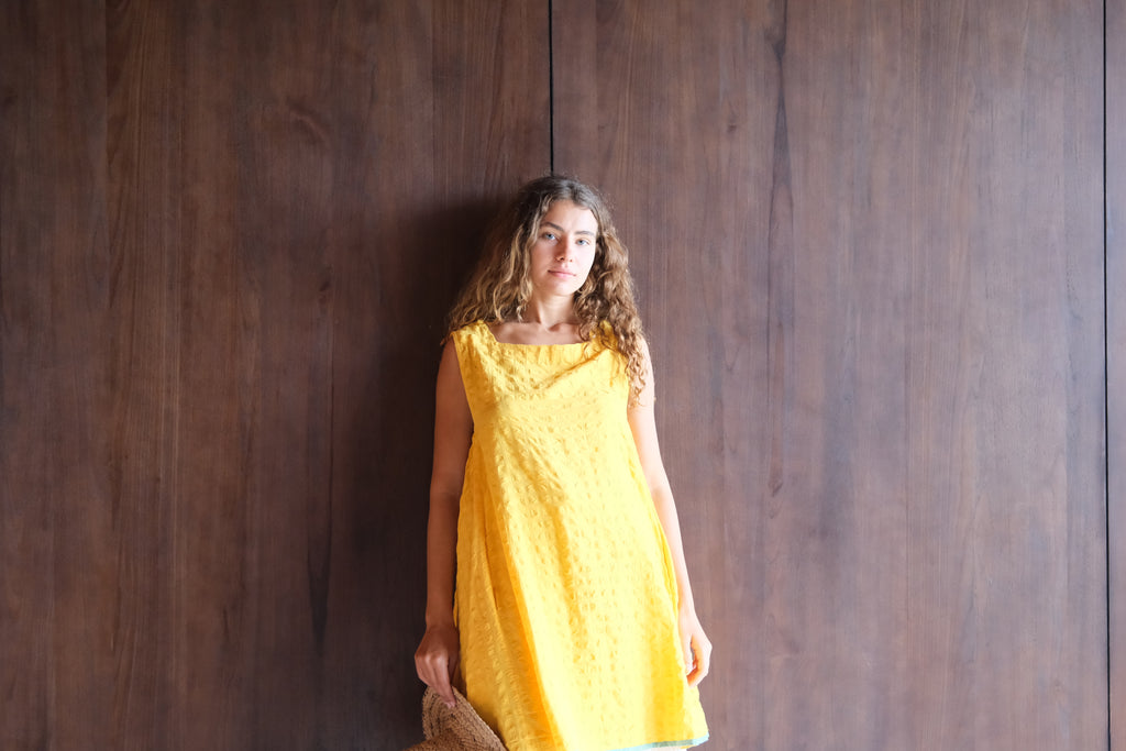 Introducing the Yellow Crinkly Dress, a collaboration between Audrey E Leary and World of Crow. This vibrant yellow mini dress features a sleeveless design and is crafted from handwoven cotton with a crinkly texture. Sustainably made, it combines minimalist style with comfort for a chic and eco-friendly wardrobe staple