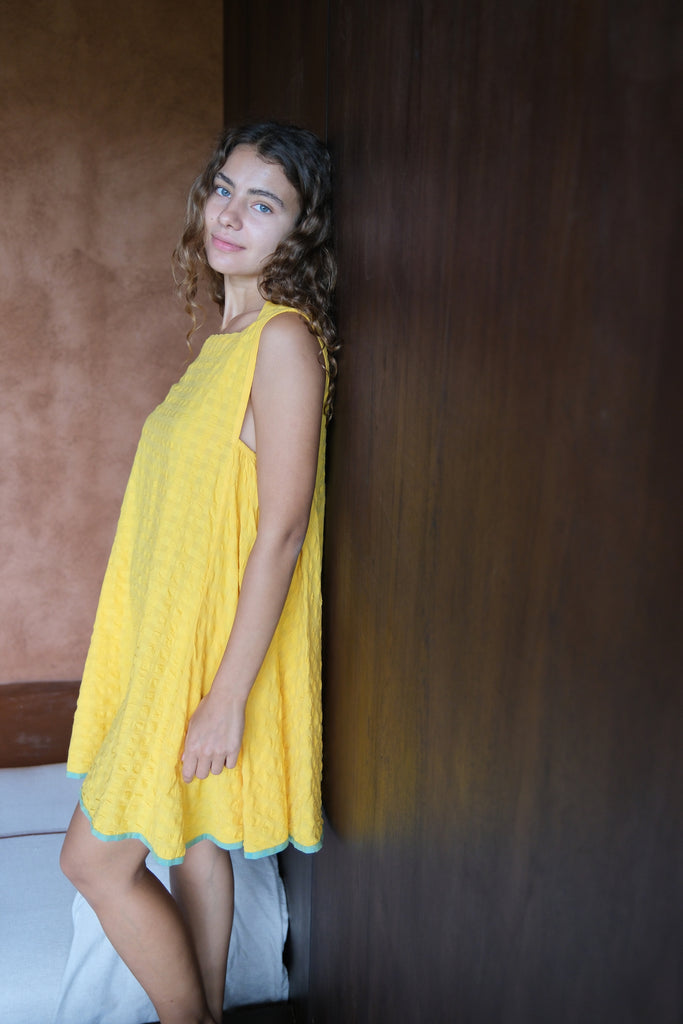 Introducing the Yellow Crinkly Dress, a collaboration between Audrey E Leary and World of Crow. This vibrant yellow mini dress features a sleeveless design and is crafted from handwoven cotton with a crinkly texture. Sustainably made, it combines minimalist style with comfort for a chic and eco-friendly wardrobe staple