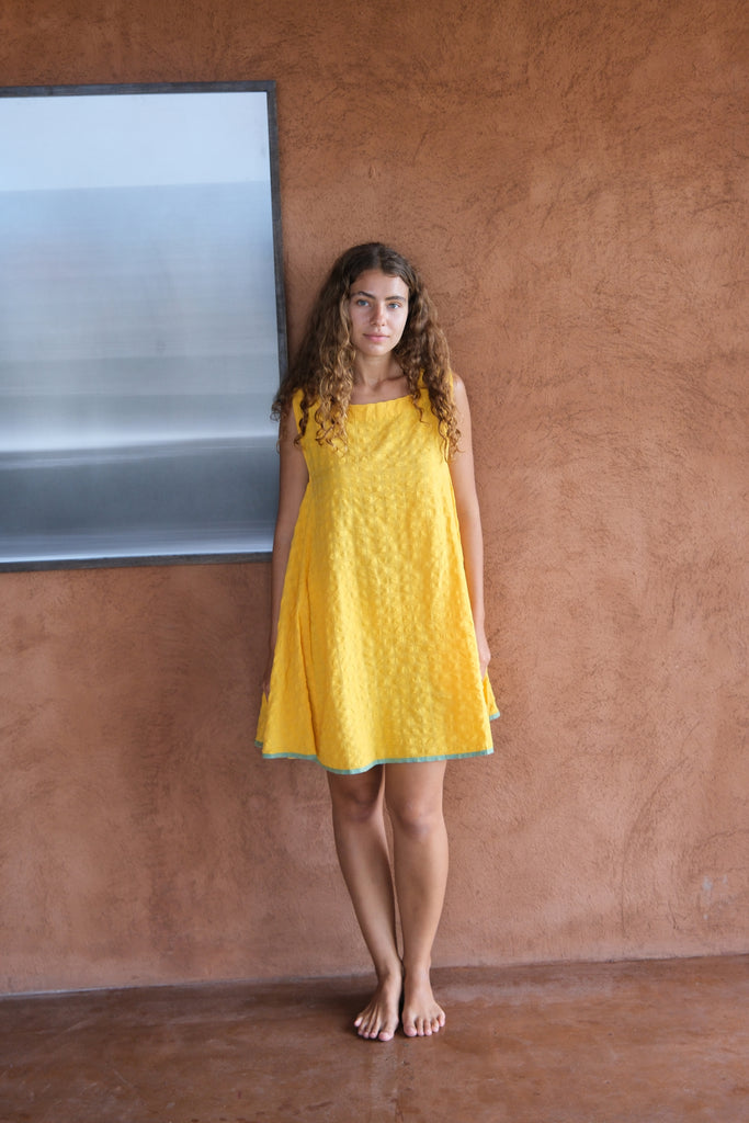 Introducing the Yellow Crinkly Dress, a collaboration between Audrey E Leary and World of Crow. This vibrant yellow mini dress features a sleeveless design and is crafted from handwoven cotton with a crinkly texture. Sustainably made, it combines minimalist style with comfort for a chic and eco-friendly wardrobe staple