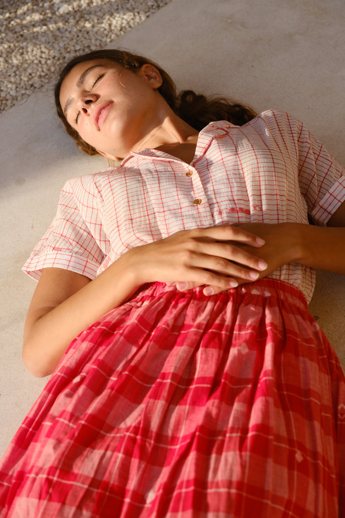 Introducing the Crimson Checkered Cuban Shirt, a collaboration between Audrey E Leary and World of Crow. This hand-woven jamdani fabric shirt is light pink with red motifs, short sleeves, and is super light and breathable, perfect for everyday wear. Sustainably made from soft cotton, paired with a red checkered skirt.