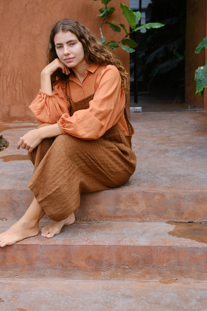 Introducing the Work-boy's Dungarees, crafted from handwoven cotton with retro self-checks and beautiful Pockets from the Audrey X Crow Collection. A women in an orange shirt and Brown work-boy's dungarees seated on steps is pictured here. These dungarees exemplify timeless craftsmanship and vintage charm, seamlessly blending comfort with style