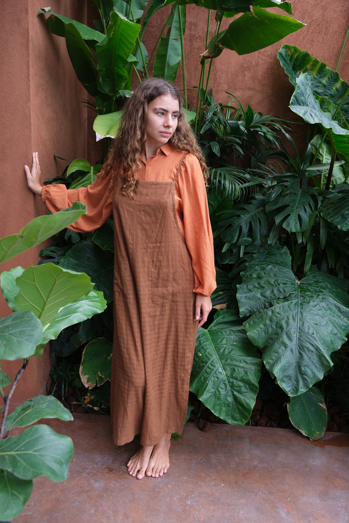 Introducing the Work-boy's Dungarees, crafted from handwoven cotton with retro self-checks and beautiful Pockets from the Audrey X Crow Collection. A women in an orange shirt and Brown work-boy's dungarees seated on steps is pictured here. These dungarees exemplify timeless craftsmanship and vintage charm, seamlessly blending comfort with style